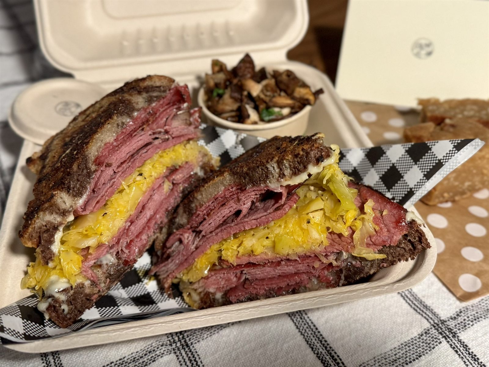 Pastrami sandwich with mustard in a takeout container, beside roasted mushrooms