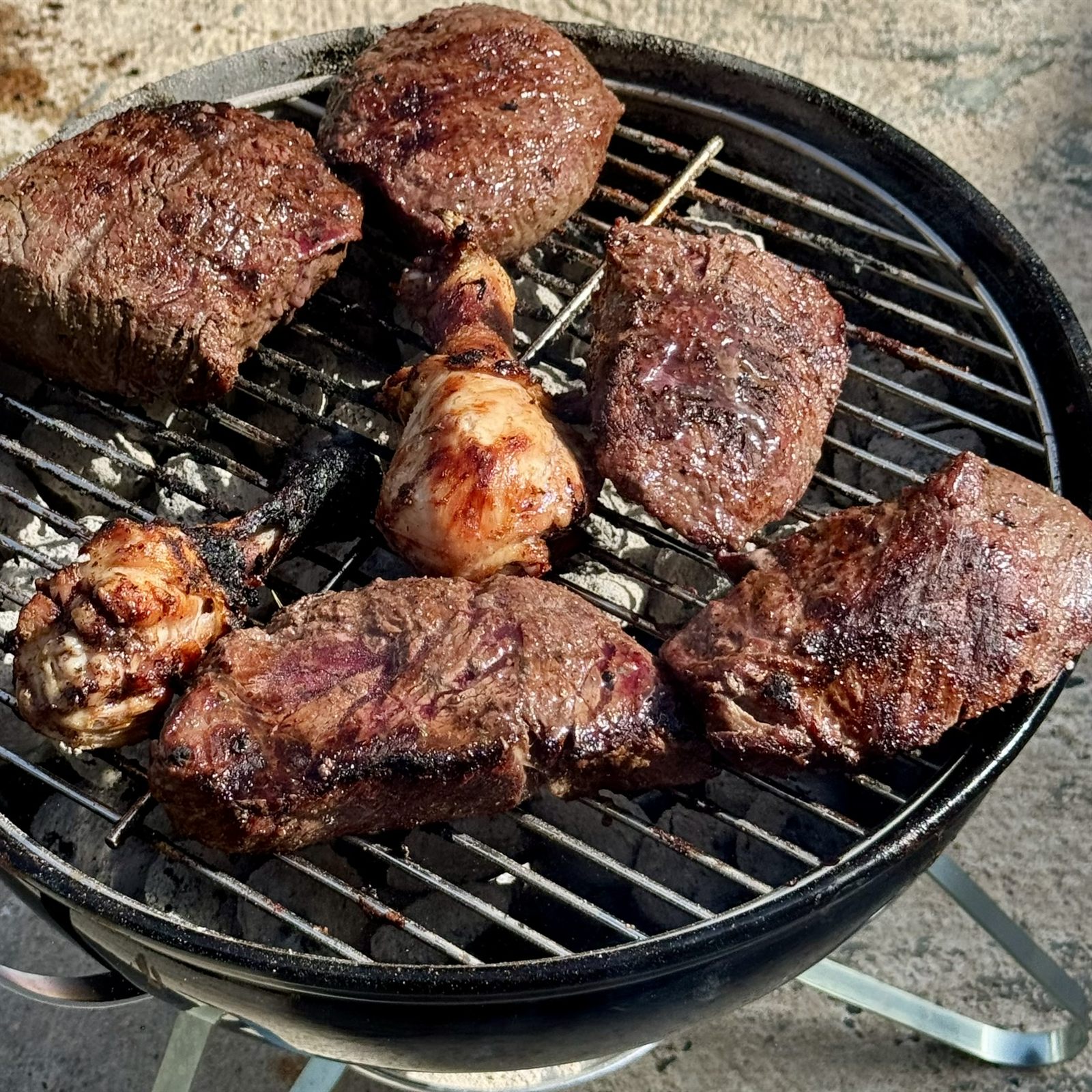 Steaks and chicken drumsticks grilling over charcoal