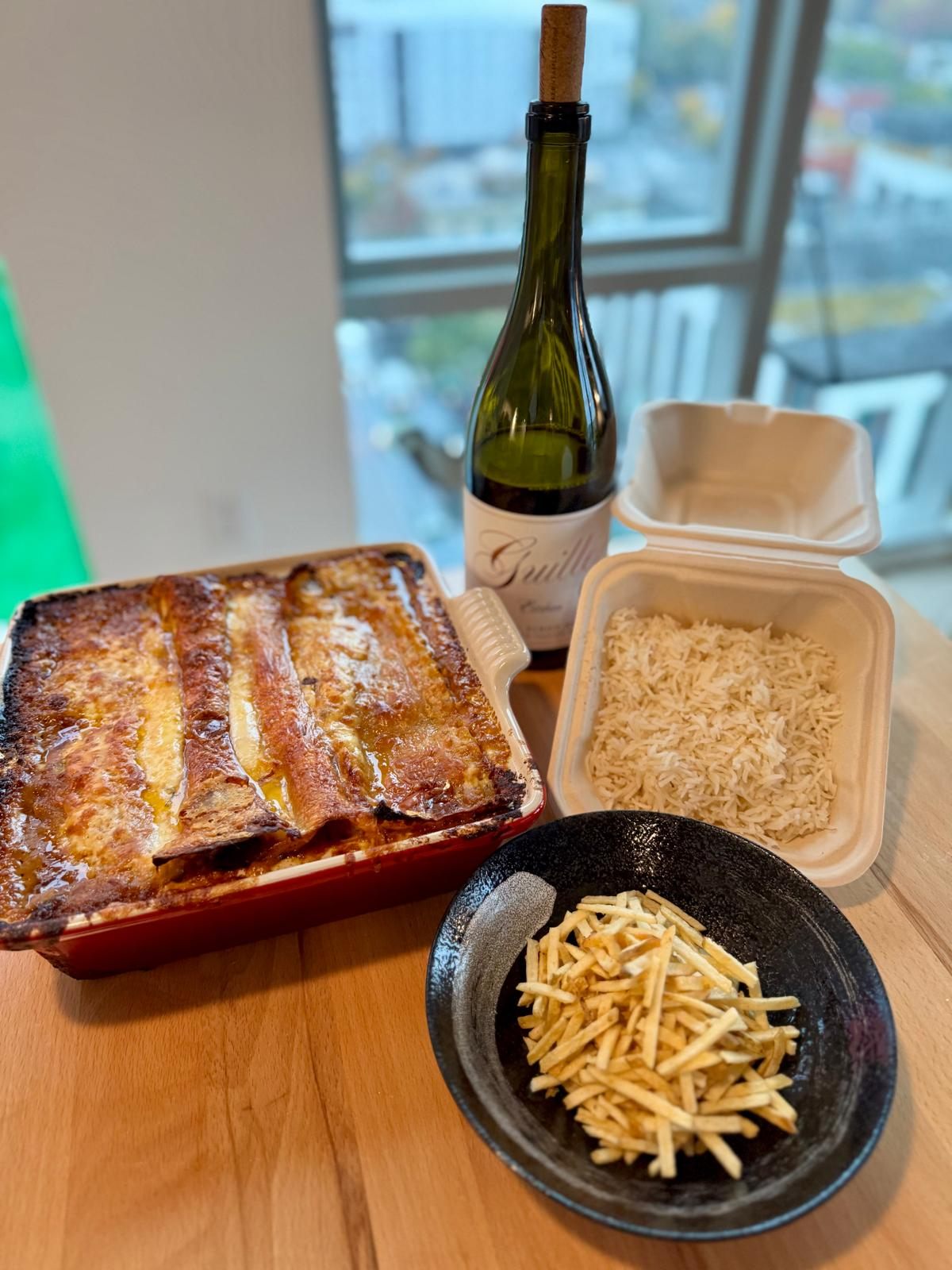 Baked lasagna, rice, a bottle of wine and shoestring potatoes laid out on a kitchen counter