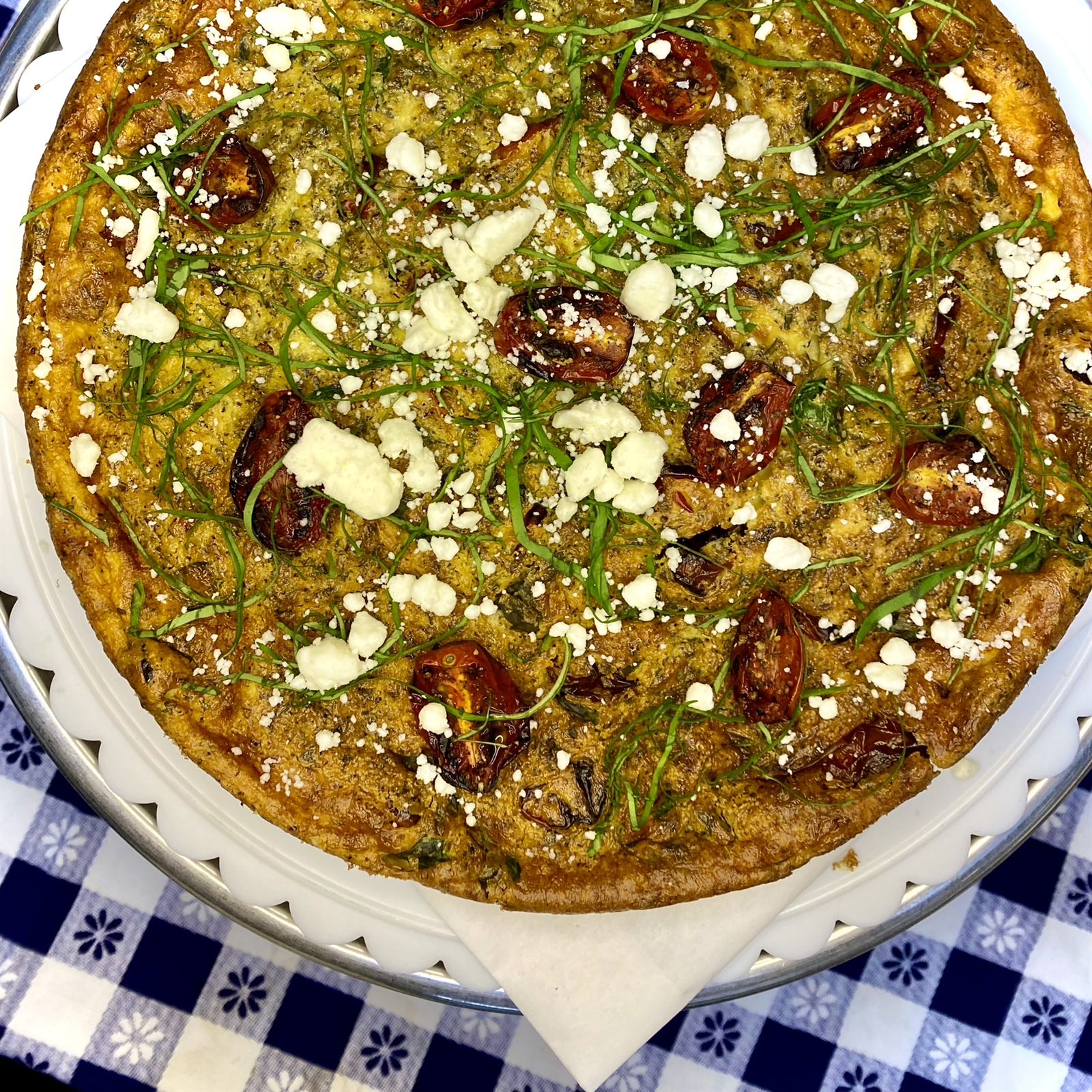Large baked frittata topped with dark fruit, crumbled cheese and herbs on a checkered cloth